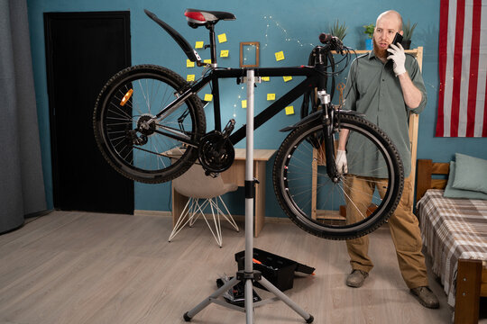 Bearded man calling on cell phone talking to mechanic asking for help in repairing broken bicycle at home