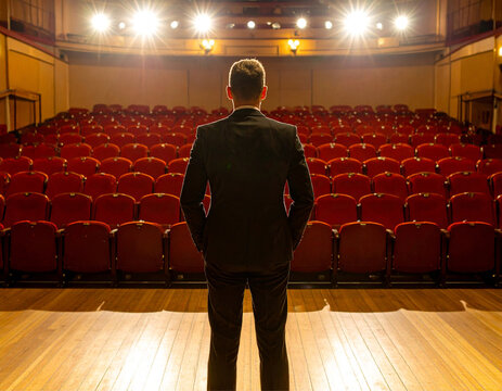 Um homem bem vestido &agrave; frente de um teatro vazio, bem iluminado, com muitas poltronas vermelhas.