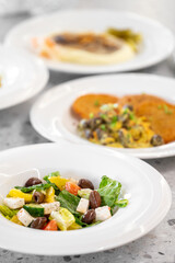 Top view of assorted Mediterranean dishes on white plates, featuring a fresh Greek salad with feta, olives, and vegetables, served on a