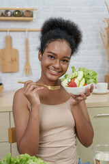 Teenage multiracial girl smiling with salad bowl in kitchen, holding fork and looking cheerful,...