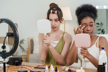 black teenage girl applying eyeshadow in front of mirror while asian teenage girl holding product...