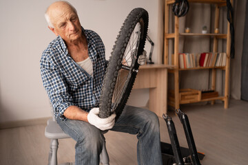 Elderly mechanic repairing bicycle holding cycle wheel in hands sitting on chair in living room....
