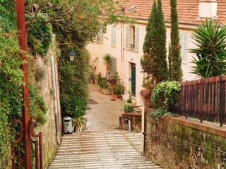 Charming narrow street in Cannes, lined with lush greenery and rustic buildings. Stone steps lead down to a cozy courtyard, creating a picturesque and inviting atmosphere.