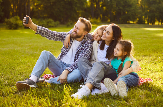 Happy family sitting together on green summer grass taking selfie using mobile phone, hugging sharing close moment, enjoying spending time together outdoors, parents and children weekend holiday