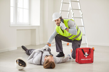Construction worker wearing safety vest and helmet kneeling beside injured coworker lying...