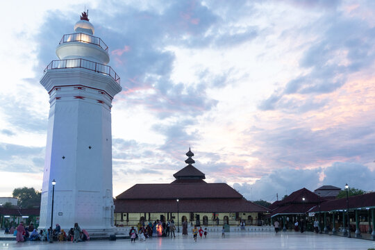 Serang, Indonesia-March 9, 2021: The iconic white lighthouse-shaped minaret and a traditional mosque building at Masjid Agung Banten Lama under a cloudy twilight sky, with people gathering.