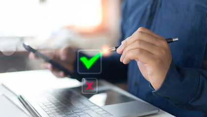Businessman holding pen and smartphone with check mark and cross icon on laptop, symbolizing...