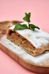Apple strudel on wooden platter with mint garnish and pink background displaying powdered sugar