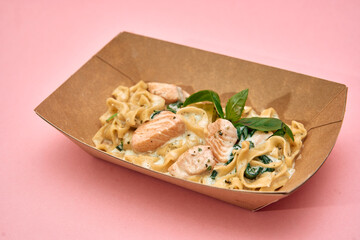 Creamy salmon pasta with spinach in eco-friendly box on pink background