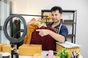 A man is live streaming his shirt on his mobile phone. On the table are boxes and shipping...