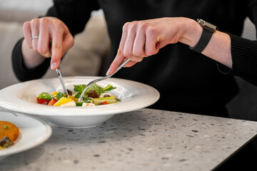 lose-up of hands holding fork and knife, eating fresh vegetable salad in a white bowl on a speckled countertop. Healthy lifestyle, casual dining concept.