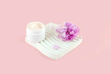 close-up of collagen cream in an open glass jar and a purple rose flower on a plaster tray on a pink background