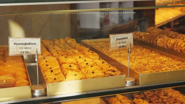 Golden mini baklava and mini saragli pastries soaked in syrup with chopped nuts, displayed in glass case inside bakery in Athens, Greece. No people 