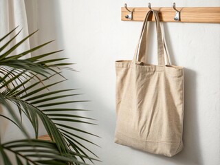 Neutral canvas tote bag hanging on wooden hook, white wall background, soft palm leaf shadow, clean minimalist setup.