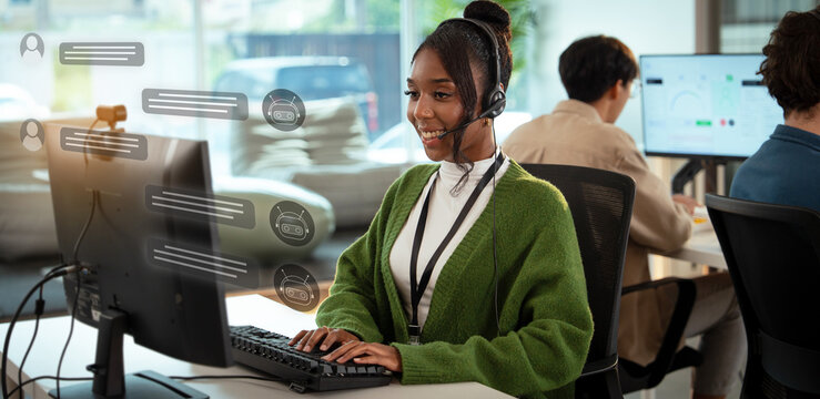 Cheerful customer support representative wearing headset is working at computer in modern office environment. background shows other team members collaborating in bright, open space - Powered by Adobe