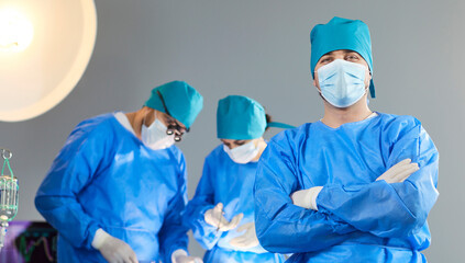 Portrait of confident surgeon wearing blue scrubs and mask standing with arms crossed in operating...