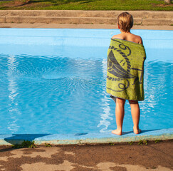 Wet child in a towel standing at the edge of a wading pool watching the vortex of water drain out.