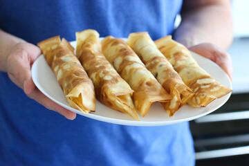 Man in blue shirt holding white plate with five rolled crepes filled with cheese. Focus on hands...