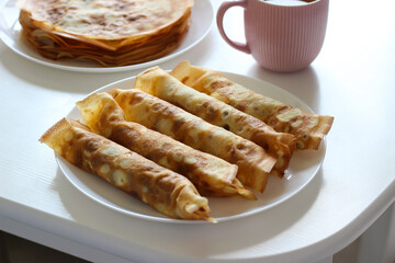 Rolled crepes with cottage cheese filling served on white plate. Pink mug in soft focus background. Cozy breakfast or light dessert idea