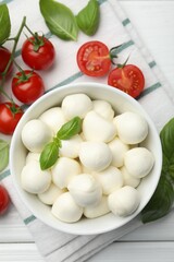 Tasty mozzarella cheese balls in bowl, basil and tomatoes on white wooden table, flat lay