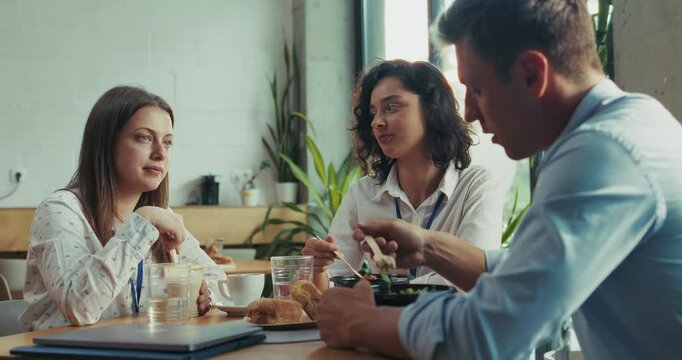 Three caucasian intern teammates enjoy lunch together in slow motion at bright sunny office cafeteria