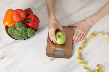 Woman weighting apple on kitchen scale at white marble table, top view