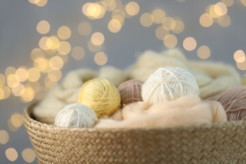 Soft balls of yarn and felting wool in wicker basket against grey background with blurred lights, closeup