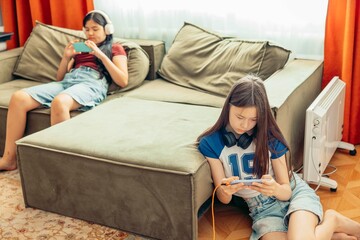 Obraz premium Asian girls relax in cozy room using smartphones with orange curtains, technology education leisure, power outlet with charging cables, video game