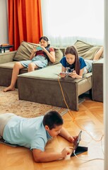 blurred boy lying on wooden floor playing with tablet while two girls relax on sofa using smartphones with orange curtains and daylight, technology education leisure, vertical