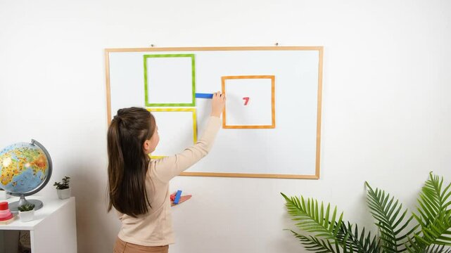 A young girl solves a math problem on a magnetic whiteboard using colorful square frames and number magnets. Shot in a cozy classroom with educational decor. 4K resolution.