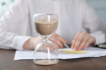 Deadline. Woman with calendar, sticky note and pen at wooden table in office, focus on hourglass