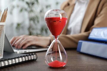 Deadline. Woman with laptop at wooden table in office, focus on hourglass