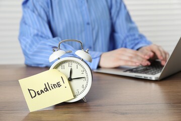 Deadline. Woman with laptop working at wooden table in office, focus on alarm clock and sticky note