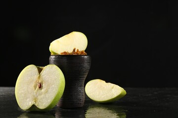 Hookah bowl with apple pieces and smoke on black table, closeup. Space for text
