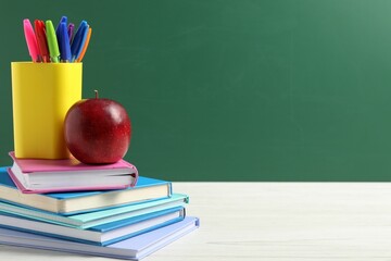Different school stationery and red apple on white wooden table against blackboard, space for text