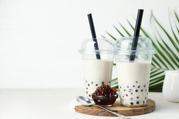 Tasty milk bubble tea in plastic cups, tapioca pearls and green leaves on beige table, space for text