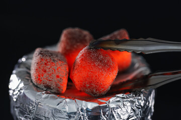 Putting smoldering charcoal cube onto hookah holder covered with foil on black background, closeup