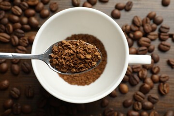 Spoon with instant coffee above cup and beans on wooden table, top view