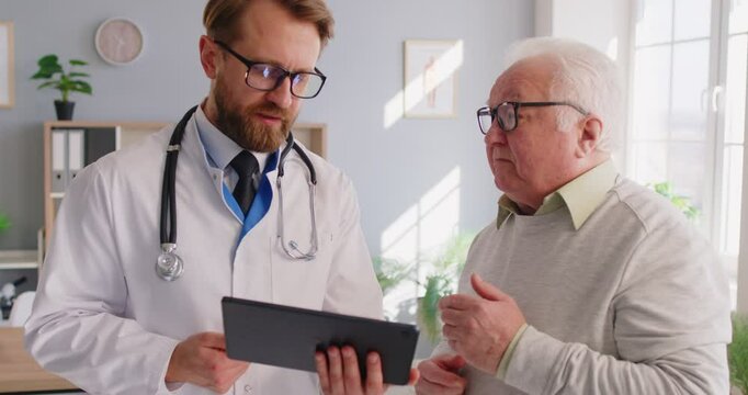 Doctor showing medical test results to senior man, explaining prescription using digital tablet app. Elderly patient visits a doctor in modern clinic. Elderly people healthcare tech concept.