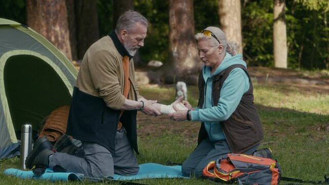 Full shot of mature Caucasian female hiker bandaging wrist of his husband sitting on grass near camping tent on chilly summer day after being injured during long hiking in wild woods