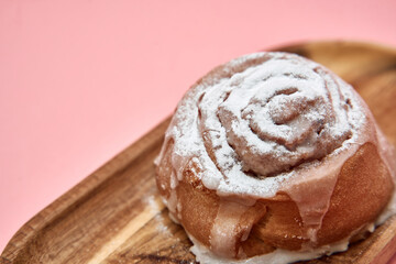 Cinnamon roll with frosting on wooden plate against pink background