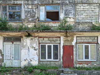 Abandoned building in ruins with peeling paint, crumbling plaster and cracks. Overgrown shrubs and grasses from neglected walls and weathered rafters. Broken windows and exposed bricks reveals decay