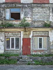 Abandoned building in ruins with peeling paint, crumbling plaster and cracks. Overgrown shrubs and grasses from neglected walls and weathered rafters. Broken windows and exposed bricks reveals decay