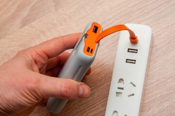 A hand connects a gray power bank to a white power strip with an orange cable for charging.