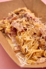 Creamy carbonara pasta with parmesan and black pepper in a takeaway box on pink background