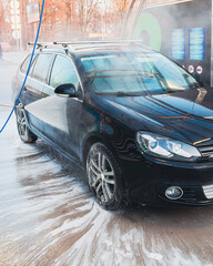 A black car gets a thorough cleaning at a car wash, with soap and water spraying everywhere.