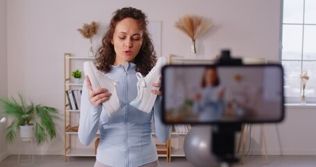 Sporty smiling young woman fitness instructor recording video and showing sports sneakers. Girl sport blogger making video for fitness blog on her smartphone about sportswear. 4k video. - Powered by Adobe
