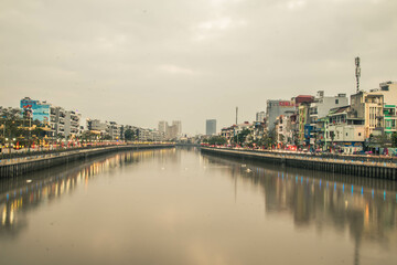Fototapeta premium The Tam Bac riverside promenade is alive with visitors enjoying the cool evening. City buildings line the bank, reflecting off the calm water as dusk approaches in Hai Phong city, Vietnam