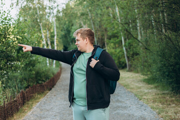 One Caucasian male hiking in the woods in summer. Travel, active lifestyle, people and hobby concept. Blonde male in sport clothes pointing finger outdoors. Traveler enjoys eco-tourism.