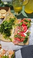 Italian sandwich with tomato, mozzarella, prosciutto and arugula on picnic table with lemonade and crackers. Concept of fresh summer lunch, healthy food and outdoor enjoyment